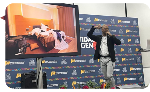 Adrián González ofreciendo una charla frente a una audiencia, mientras muestra una imagen de una habitación desordenada en la pantalla grande detrás de él.