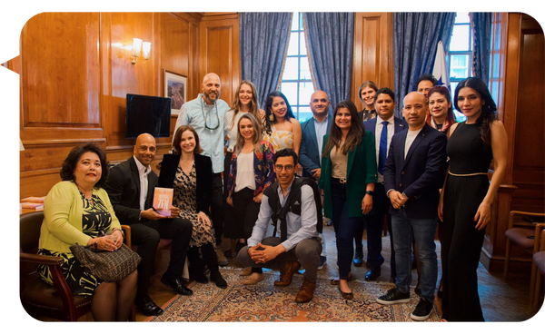 Grupo de personas sonrientes posando en una sala elegante de madera, con cortinas azul oscuro y luz natural. Adrián González se encuentra de pie, vestido con camisa clara y chaqueta, rodeado de compañeros.