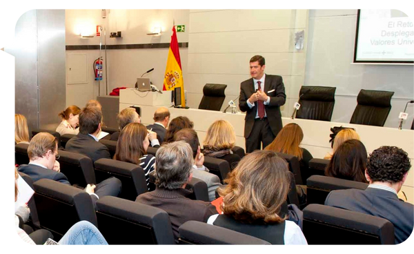 Borja Milans de Bosch dando una conferencia en una sala con varios asistentes sentados. Bandera de España en el fondo.