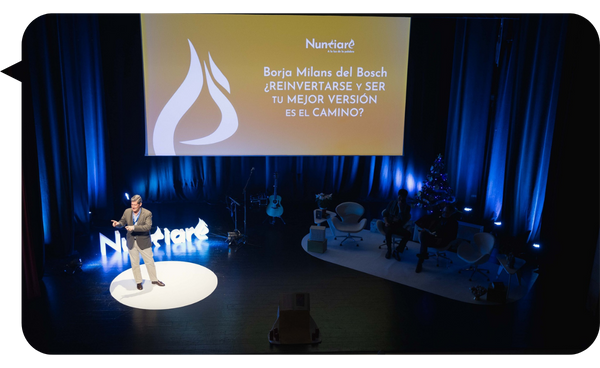 Borja Milans de Bosch presentando en un escenario iluminado, con una pantalla de fondo que muestra el tema de su charla sobre reinvención personal.