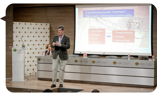 Borja Milans del Bosch dando una conferencia en una universidad, junto a una presentación sobre las dimensiones existenciales del ser humano.