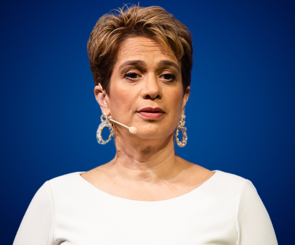 Janine Sikaffy, conferencista y mentora, hablando en un escenario con fondo azul, vestida de blanco y usando un micrófono de diadema.