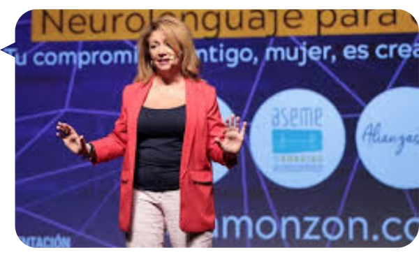 Lidia Monzón en una conferencia sobre neurolenguaje, de pie en el escenario con un fondo azul y logotipos de patrocinadores, vestida con un blazer rojo y camiseta negra.