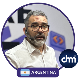 Martín Quirós hablando en una conferencia, con auriculares y fondo corporativo, junto a un logo de Argentina y de la organización CHM.