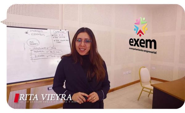 Rita Vieyra frente a una pizarra durante una sesión de capacitación empresarial, con el logo de EXEM Acompañamiento Empresarial al fondo.