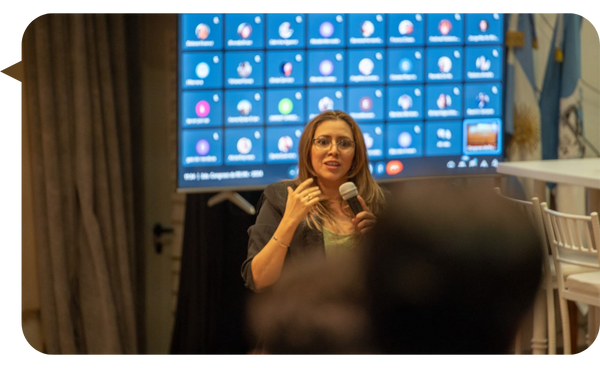 Rita Vieyra dando una conferencia con micrófono en mano, frente a una pantalla con participantes en una videollamada.