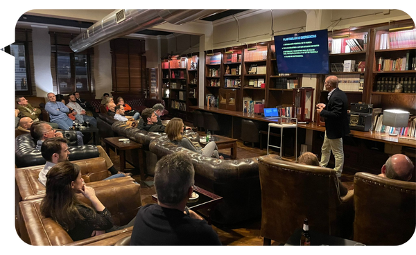 Manolo Cámara dando una conferencia en una sala estilo biblioteca, con una audiencia sentada en sofás.