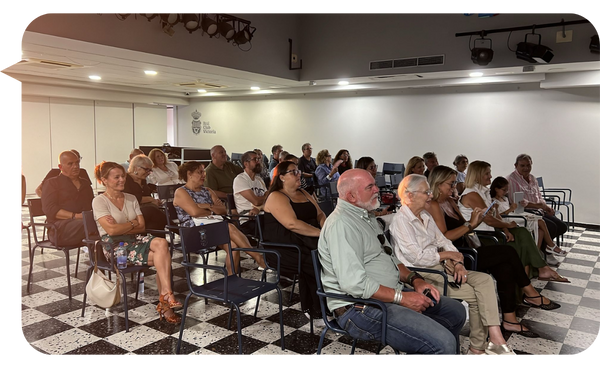 Audiencia atenta en una conferencia, sentados en sillas en un salón con suelo blanco y negro.