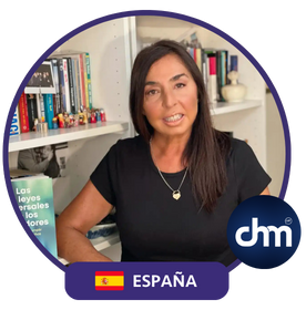 Mujer sonriente, con cabello largo y oscuro, vestida con camiseta negra, sentada frente a una estantería con libros, junto a un banner de CHM y una bandera de España.