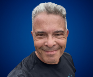 Adolfo Alvarez León sonriente con camiseta deportiva negra, fondo azul.