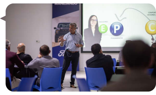 Alejandro Delobelle impartiendo una conferencia a un grupo de profesionales, con una presentación sobre emociones, pensamientos y acciones proyectada en la pantalla.