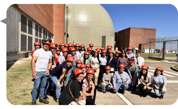 Grupo de personas con cascos de seguridad naranjas posando frente a instalaciones industriales al aire libre.