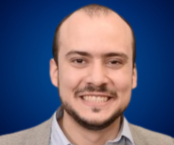 Retrato de Daniel Crespo, conferencista, con fondo azul, vistiendo camisa clara y chaqueta formal, mostrando una sonrisa cálida"