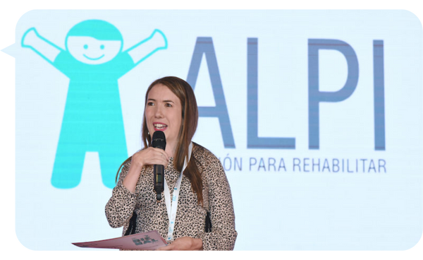 Mujer sonriente con cabello castaño sosteniendo un micrófono, vestida con una blusa estampada, de pie frente a un escenario con el logo de ALPI y la frase "Fundación para Rehabilitar".