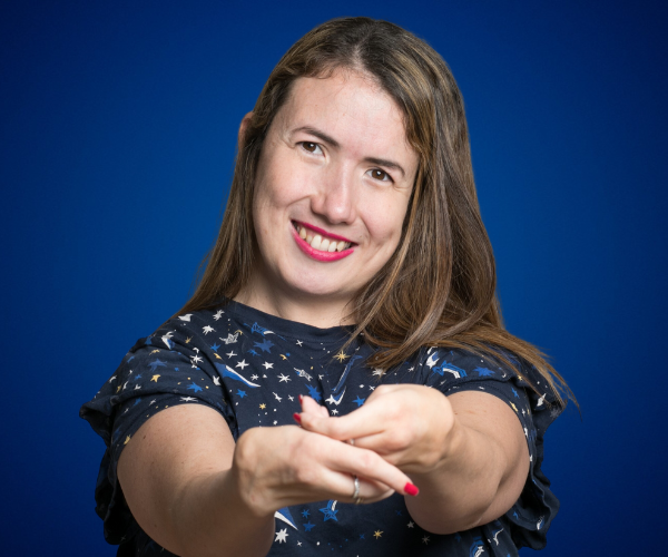 Mujer sonriente con cabello castaño, vestida con una blusa azul con estampado de estrellas, posando con las manos extendidas hacia la cámara, sobre un fondo azul intenso.