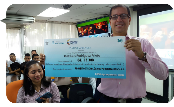 Hombre sonriente sostiene un cheque grande con un valor de 84.113.388 pesos colombianos, mientras una mujer a su lado sonríe y sostiene un celular en una sala de reuniones.