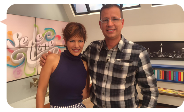 Hombre con camisa a cuadros y mujer con blusa sin mangas posan sonrientes en un set de televisión.