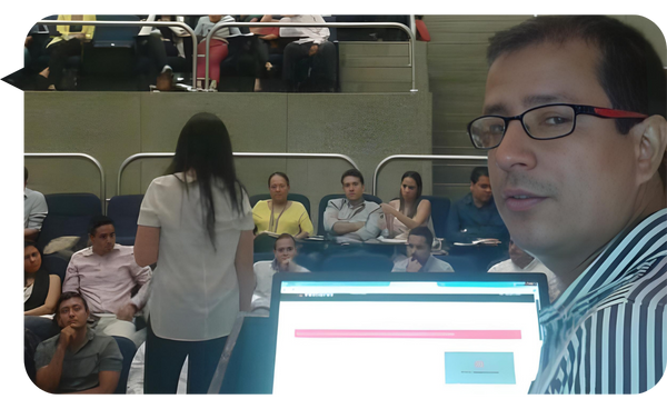 Hombre con gafas frente a una computadora portátil durante una conferencia, con asistentes atentos en una sala de conferencias.