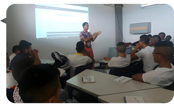 Hombre de pie frente a un proyector, dando una clase a un grupo de estudiantes en un aula.