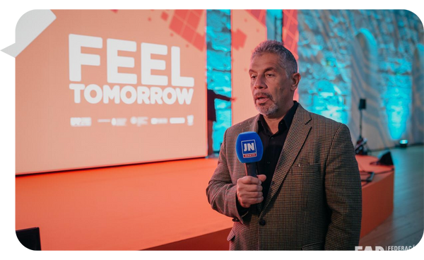 Pancho Campo siendo entrevistado con un micrófono de "JN", frente a un escenario con la proyección "Feel Tomorrow".