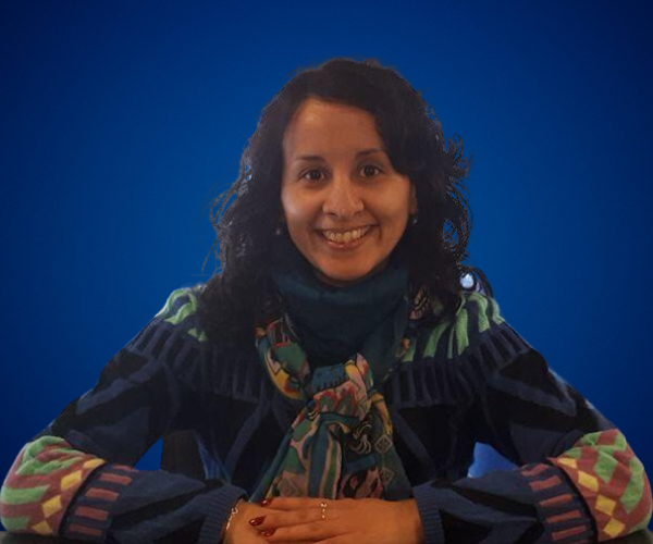 Mujer sonriente con cabello oscuro y rizado, vestida con suéter de colores y bufanda, posando frente a un fondo azul.