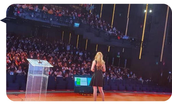 "Yami Almaguer Gil de espaldas, presentando en un auditorio lleno de asistentes con un podio y pantalla en el escenario."