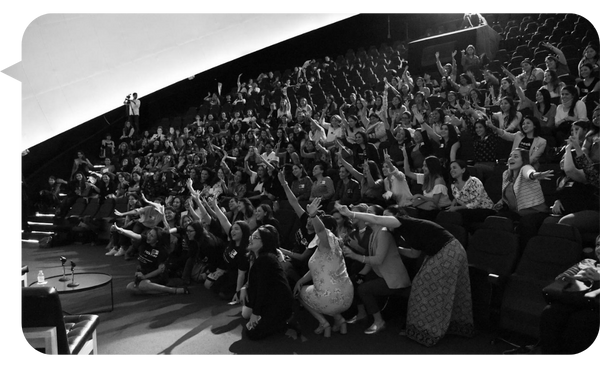 "Grupo numeroso de personas en un auditorio participando con entusiasmo durante una dinámica liderada por Yami Almaguer Gil."