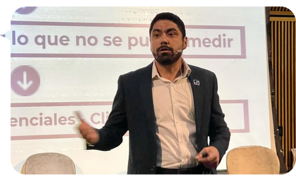 "Daniel Ortiz impartiendo una conferencia, vestido con traje formal y micrófono de diadema, frente a una pantalla con frases motivacionales."