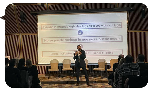 "Daniel Ortiz impartiendo una conferencia frente a una pantalla con gráficos y frases motivacionales, rodeado de una audiencia en un auditorio moderno."