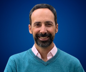 Retrato de Luis Felipe Avella Villegas sonriendo, vistiendo un suéter azul y camisa rosada, sobre un fondo azul oscuro.