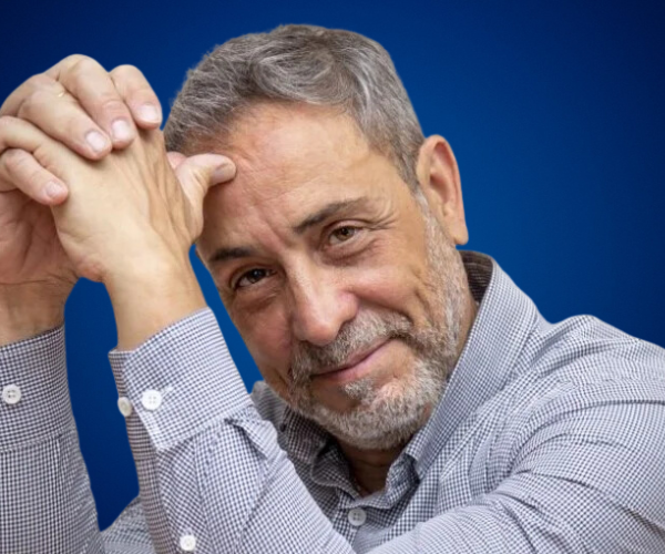 Retrato de Marcelo Carbone, conferencista internacional, mirando directamente a la cámara con una expresión amable y las manos entrelazadas frente a un fondo azul.