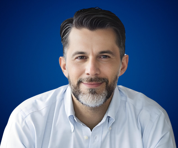 Allan Loría, experto en longevidad social y diversidad generacional, posando con un fondo azul que resalta su profesionalismo y cercanía.