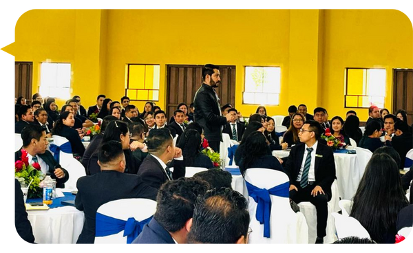 Mario de Paz interactuando con una audiencia en un evento, rodeado de asistentes sentados en mesas decoradas, en un espacio amplio y luminoso.