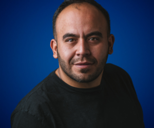 Retrato de Gaspar Contreras, hombre con barba corta y cabeza rapada, vistiendo una camiseta negra, con una expresión segura y un fondo azul sólido.