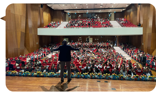 Javier Benítez Campos de espaldas, dirigiéndose a una audiencia masiva en un auditorio lleno, con un escenario decorado con flores.