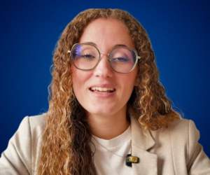 Iris Shara, conferencista y coach, con cabello rizado, gafas y vestimenta profesional clara, sonriendo mientras habla frente a un fondo azul.