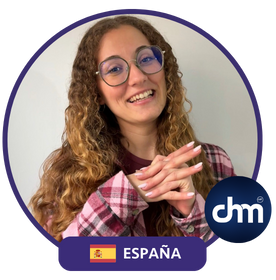 Iris Shara, conferencista y coach, sonriendo con manos entrelazadas, vistiendo una camisa a cuadros y gafas, con la bandera de España y el logo de "chm".