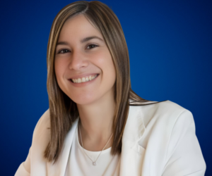 Retrato de Mónica Caruso, sonriendo con un fondo azul, vestida con un traje blanco y una cadena plateada.