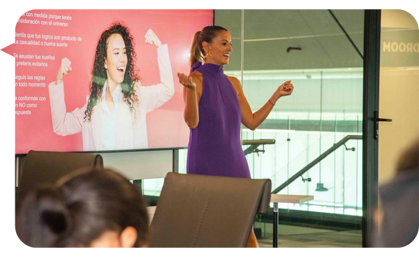 Rebeca Barzuna, vestida con un elegante vestido morado, imparte una conferencia en una sala de reuniones moderna. En la pantalla detrás de ella, se muestra una imagen motivacional de una mujer levantando los brazos con confianza.