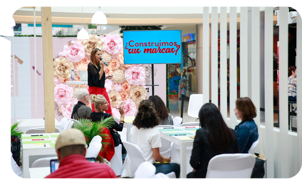 Rebeca Barzuna en una conferencia en un espacio decorado con flores y mobiliario blanco, presentando ante una audiencia atenta mientras sostiene un micrófono. En la pantalla detrás de ella se lee "¿Construimos sus marcas?".