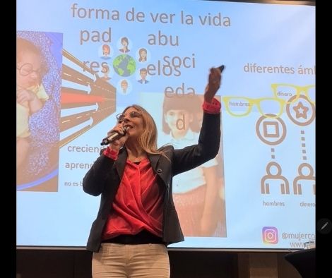 Vivianne Verschae ofreciendo una charla con micrófono en mano frente a una presentación proyectada sobre creencias y roles sociales.