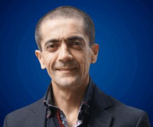 Christian Estay, conferencista experto, vistiendo saco oscuro y camisa formal, posando con una expresión amable sobre fondo azul.