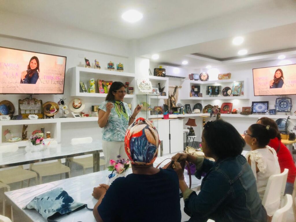 Vivianne Verschae impartiendo un taller presencial a un grupo de mujeres, rodeada de artesanías y pantallas con su imagen promocional.
