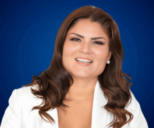 Ximena Cárdenas, conferencista profesional, con cabello suelto ondulado y chaqueta blanca, sonriente frente a un fondo azul.