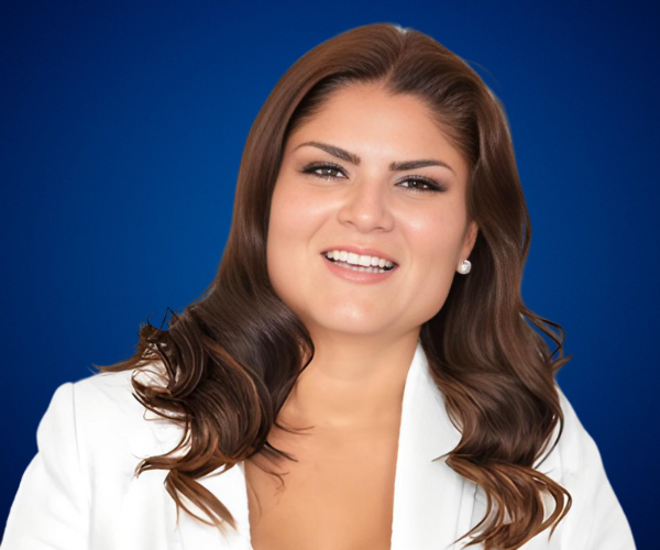 Ximena Cárdenas, conferencista profesional, con cabello suelto ondulado y chaqueta blanca, sonriente frente a un fondo azul.