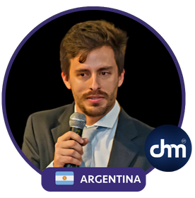 Agustín Ochoa hablando con micrófono durante una conferencia en Argentina, vestido con traje formal.