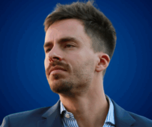 Agustín Ochoa con expresión reflexiva, vestido con blazer azul y camisa de rayas, sobre fondo azul institucional.