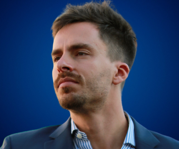 Agustín Ochoa con expresión reflexiva, vestido con blazer azul y camisa de rayas, sobre fondo azul institucional.