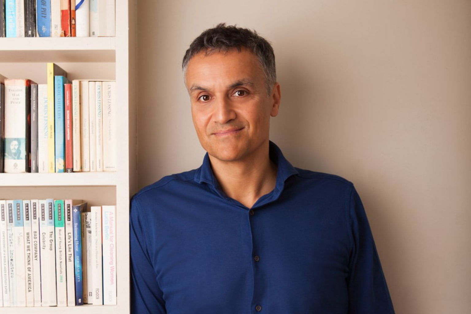 Carl Honoré de pie junto a una estantería de libros, luciendo una camisa azul, en un ambiente cálido e intelectual.