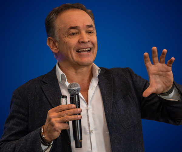 Mauricio Duarte con saco azul y camisa blanca sobre fondo azul, en imagen profesional de perfil frontal.